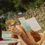 Donna con occhiali che legge un libro e tiene un bicchiere di vino bianco accanto a una piscina