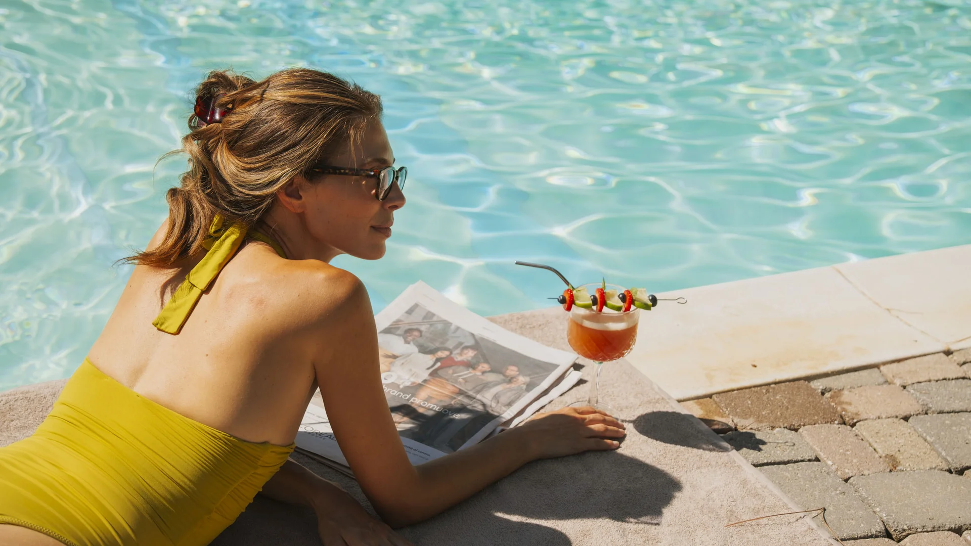 Donna in costume giallo sdraiata vicino a piscina con cocktail e giornale